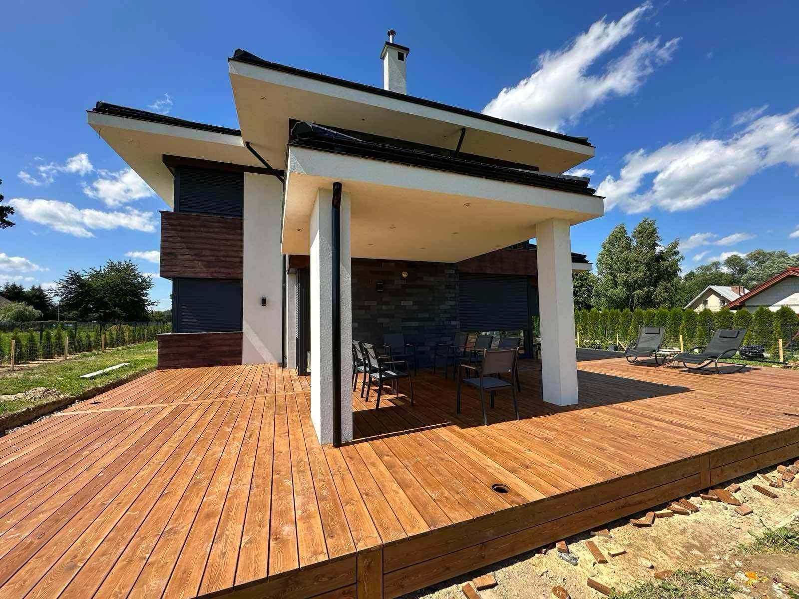 Nowoczesny dom z drewnianą elewacją i przestronnym tarasem z desek, częściowo wykończonym, z widocznymi niedokończonymi krawędziami i elementami mocującymi na obrzeżach, na tle błękitnego nieba.