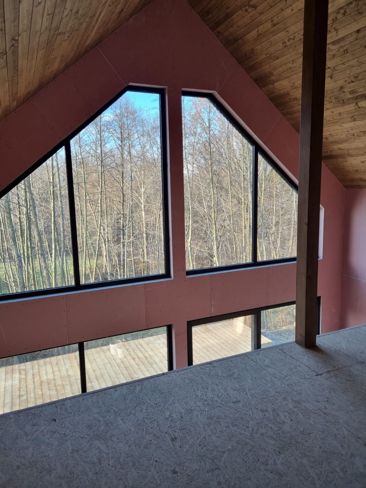 Wnętrze domu szkieletowego w trakcie budowy, widoczne okna o nietypowym kształcie i surowe drewno na podłodze i suficie.