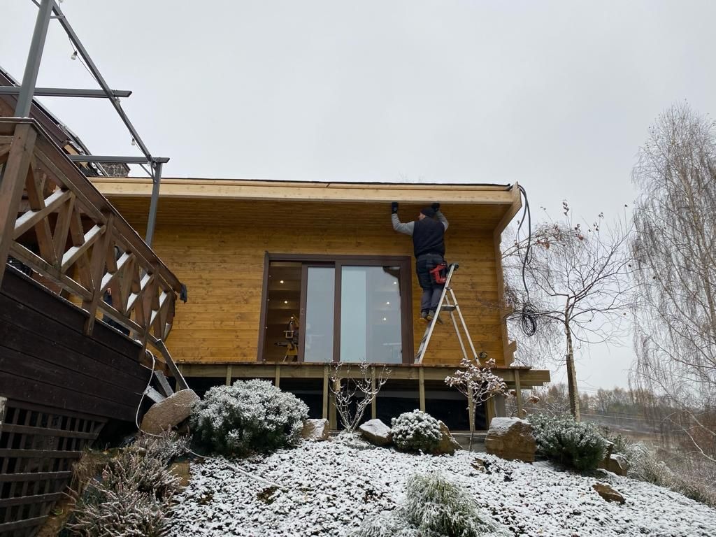 Montaż oświetlenia zewnętrznego na drewnianym domu szkieletowym w zimowej scenerii. Pracownik na drabinie instaluje przewody. Częściowo ośnieżony teren z roślinnością i elementami tarasu.
