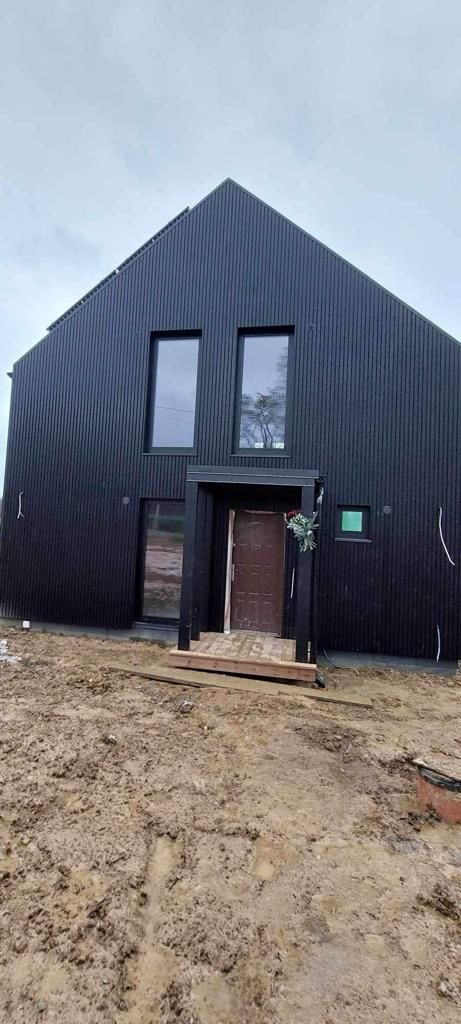 Nowoczesny, czarny dom szkieletowy z pionowym deskowaniem, widoczne okna i drzwi wejściowe z małym gankiem, teren wokół domu w trakcie prac wykończeniowych, ziemia błotnista po deszczu.