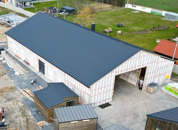 Wysoki budynek w trakcie budowy z widoczną konstrukcją szkieletową i szarą blachą na dachu, obok mniejsze budynki z podobnym dachem, widok z góry.