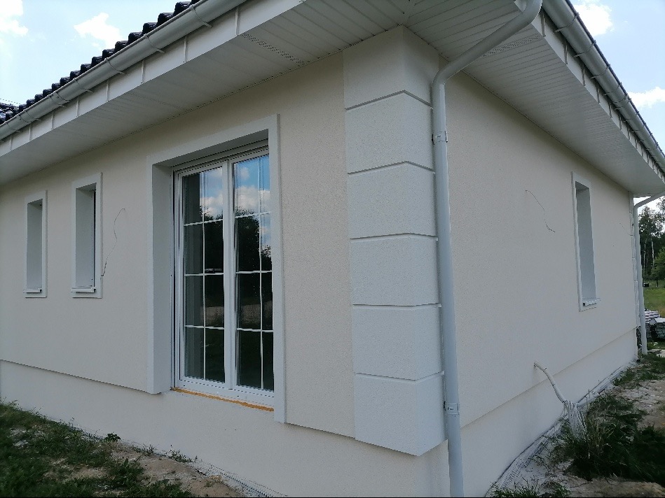 Wykończona elewacja domu jednorodzinnego z kremowym tynkiem strukturalnym, białymi obramowaniami okien i ozdobnymi narożnikami z boniami, widoczny fragment dachu i rynna.