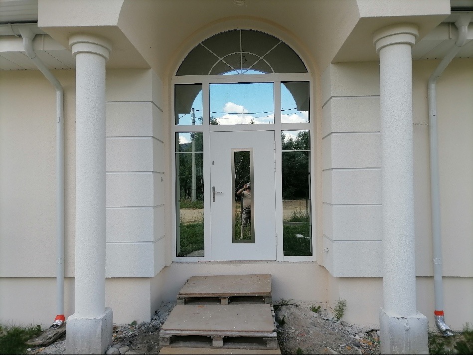 Wejście do domu z kolumnami i łukowym oknem nad drzwiami, widoczne schody z palet i odbicie fotografa w szybie.