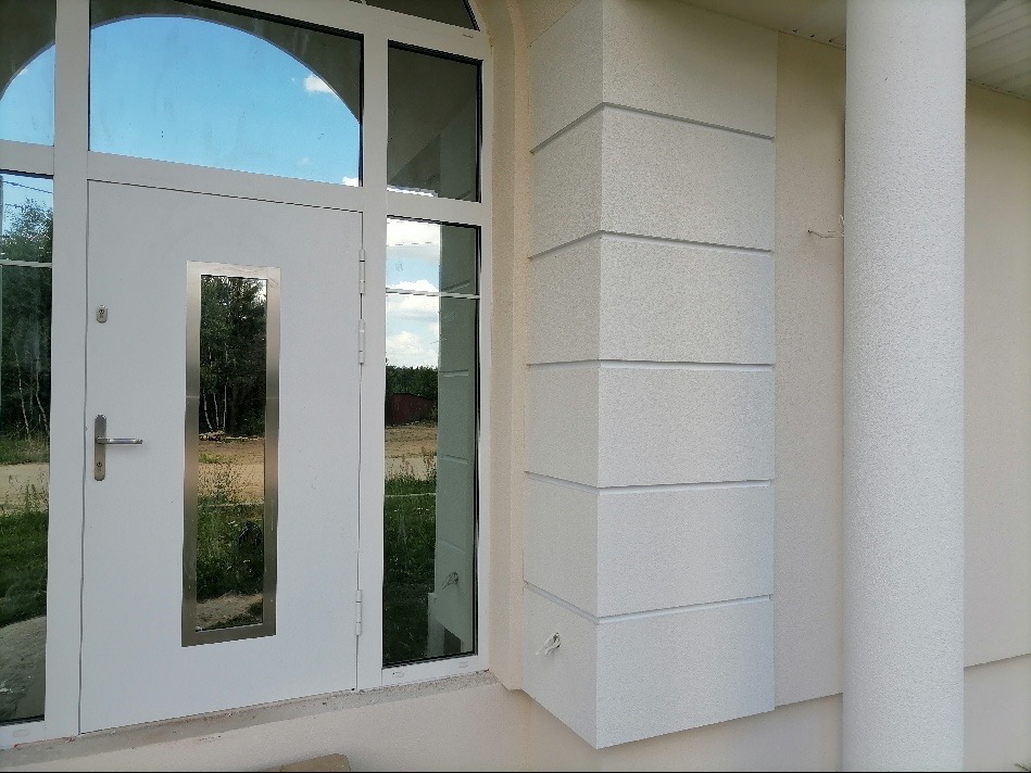 Fragment elewacji domu z białym tynkiem strukturalnym, widoczna kolumna i narożnik z dekoracyjnymi poziomymi wgłębieniami, obok białe drzwi wejściowe z przeszkleniem.