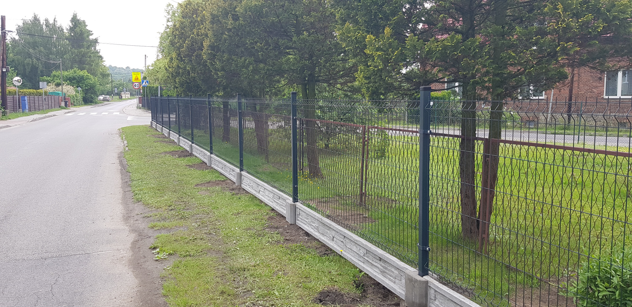 Nowoczesne, panelowe ogrodzenie z podmurówką imitującą kamień, widok z perspektywy ulicy, słupki w kolorze grafitowym, siatka w kolorze ciemnozielonym.