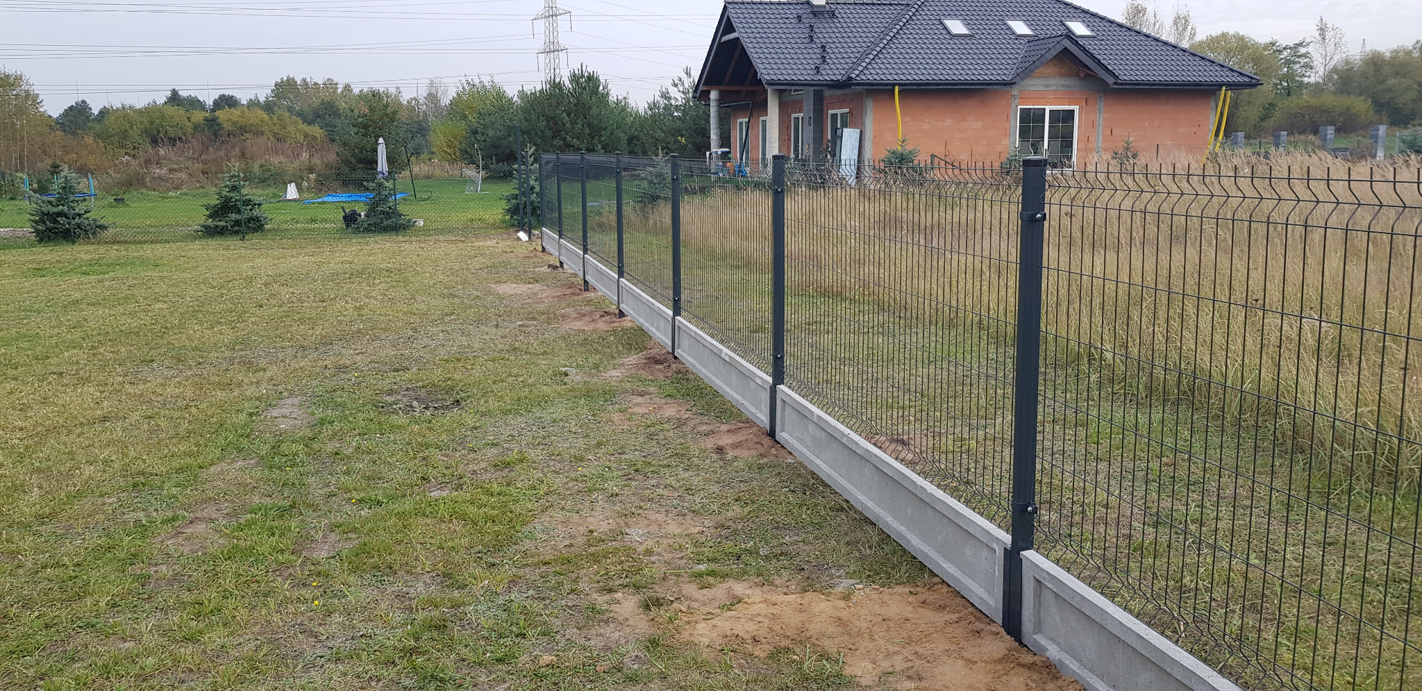 Panelowe ogrodzenie z podmurówką betonową, widoczne na tle trawnika i domu w trakcie budowy.