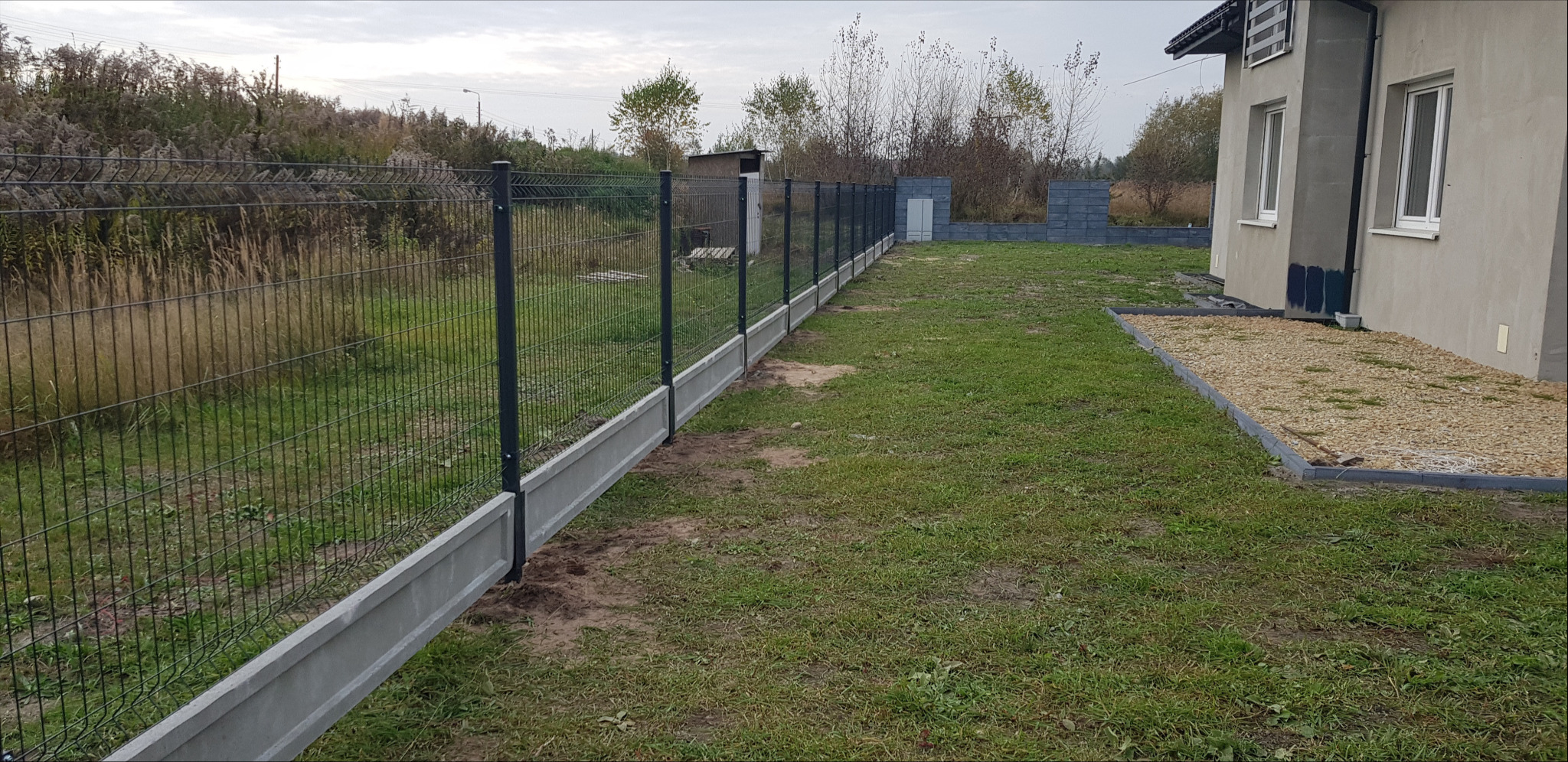 Nowoczesne ogrodzenie panelowe z podmurówką betonową, widok z perspektywy trawnika w kierunku posesji i sąsiedniego pola.