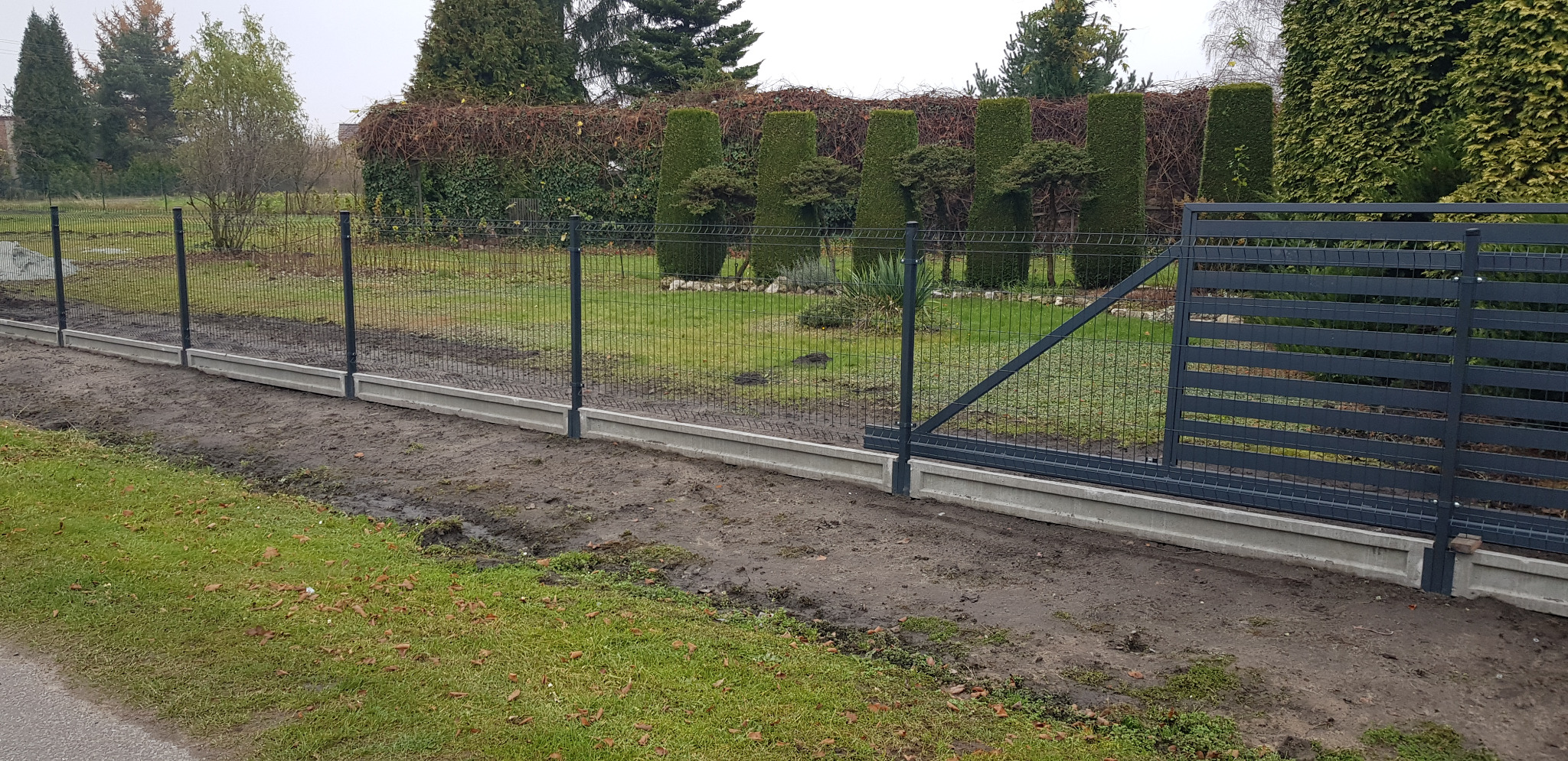 Nowoczesne ogrodzenie panelowe z podmurówką betonową i przesuwną bramą wjazdową na posesję z zielonym trawnikiem i ozdobnymi tujami w tle.