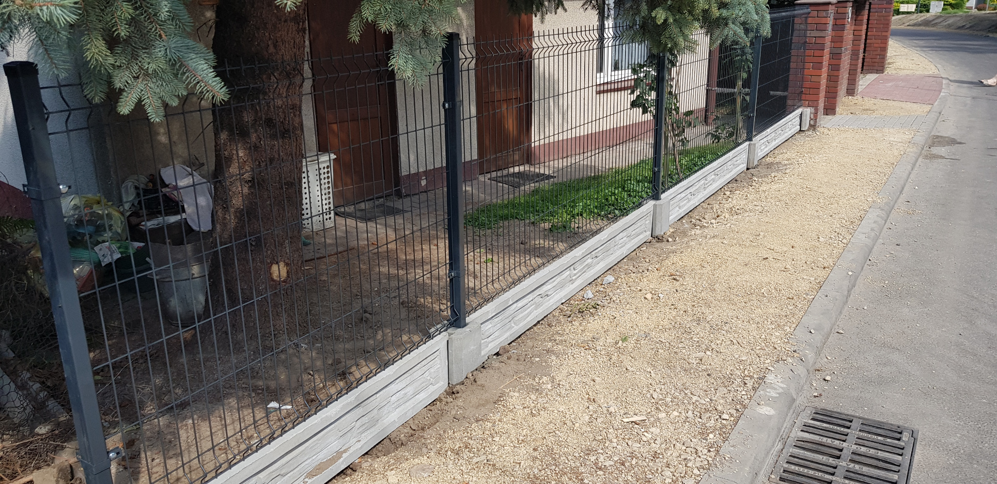 Nowoczesne ogrodzenie panelowe z podmurówką imitującą kamień, czarne słupki, widok z zewnątrz posesji, częściowo zasłonięte przez gałęzie drzewa iglastego, w tle budynek mieszkalny z brązowymi...