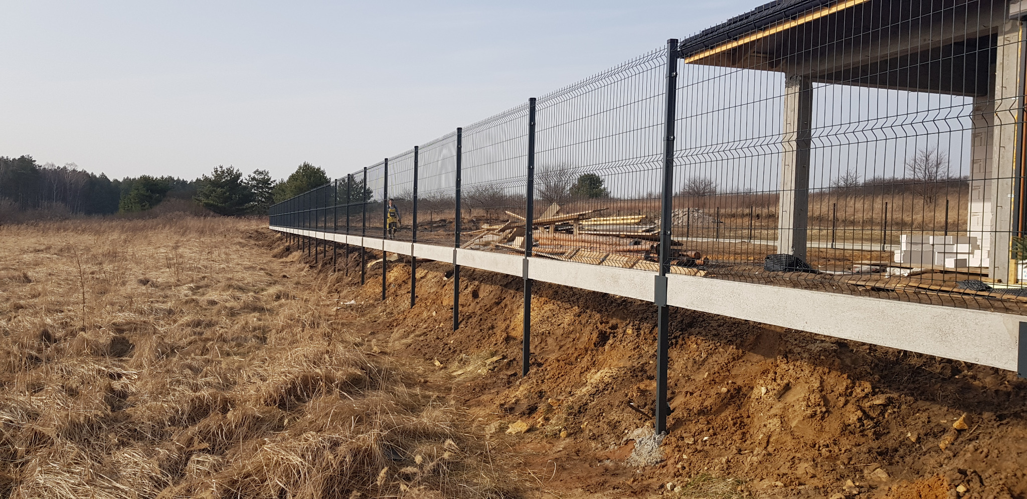 Długa, metalowa siatka ogrodzeniowa na betonowej podmurówce w trakcie instalacji, widoczne drewniane elementy konstrukcyjne i budynek w stanie surowym w tle, sucha trawa na pierwszym planie.