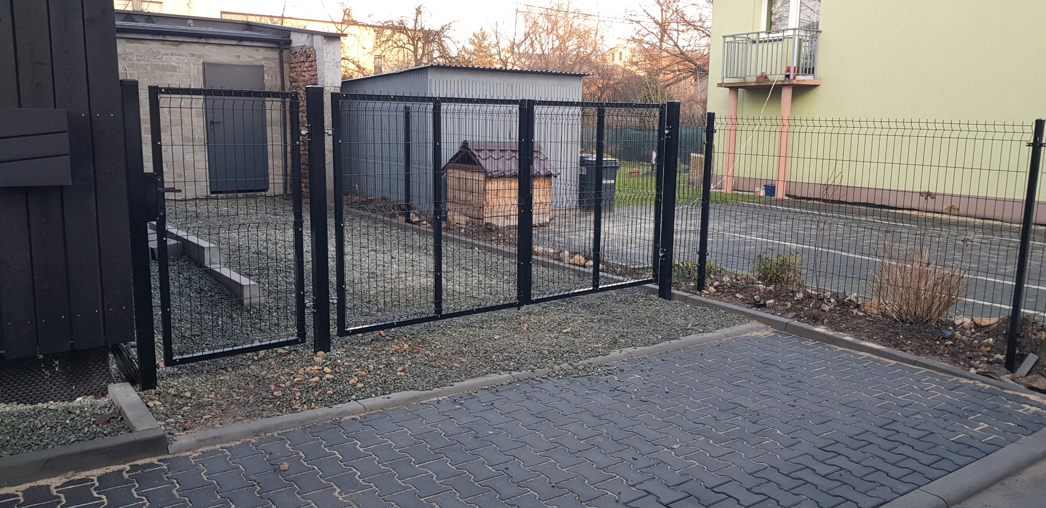 Nowoczesne, czarne ogrodzenie panelowe z bramą uchylną, widok z zewnątrz posesji, na podłożu szara kostka brukowa i drobny żwir.