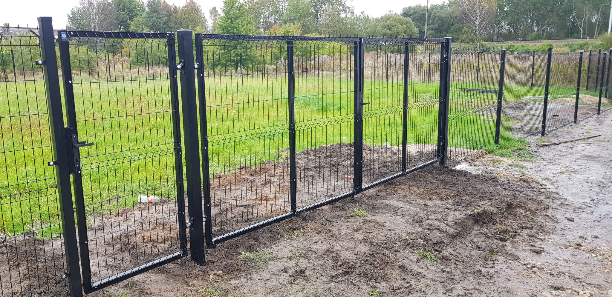 Czarna, metalowa brama panelowa z furtką i ogrodzenie systemowe z siatki, zamontowane na nierównym terenie z widoczną ziemią i trawą, w tle drzewa i tory kolejowe.