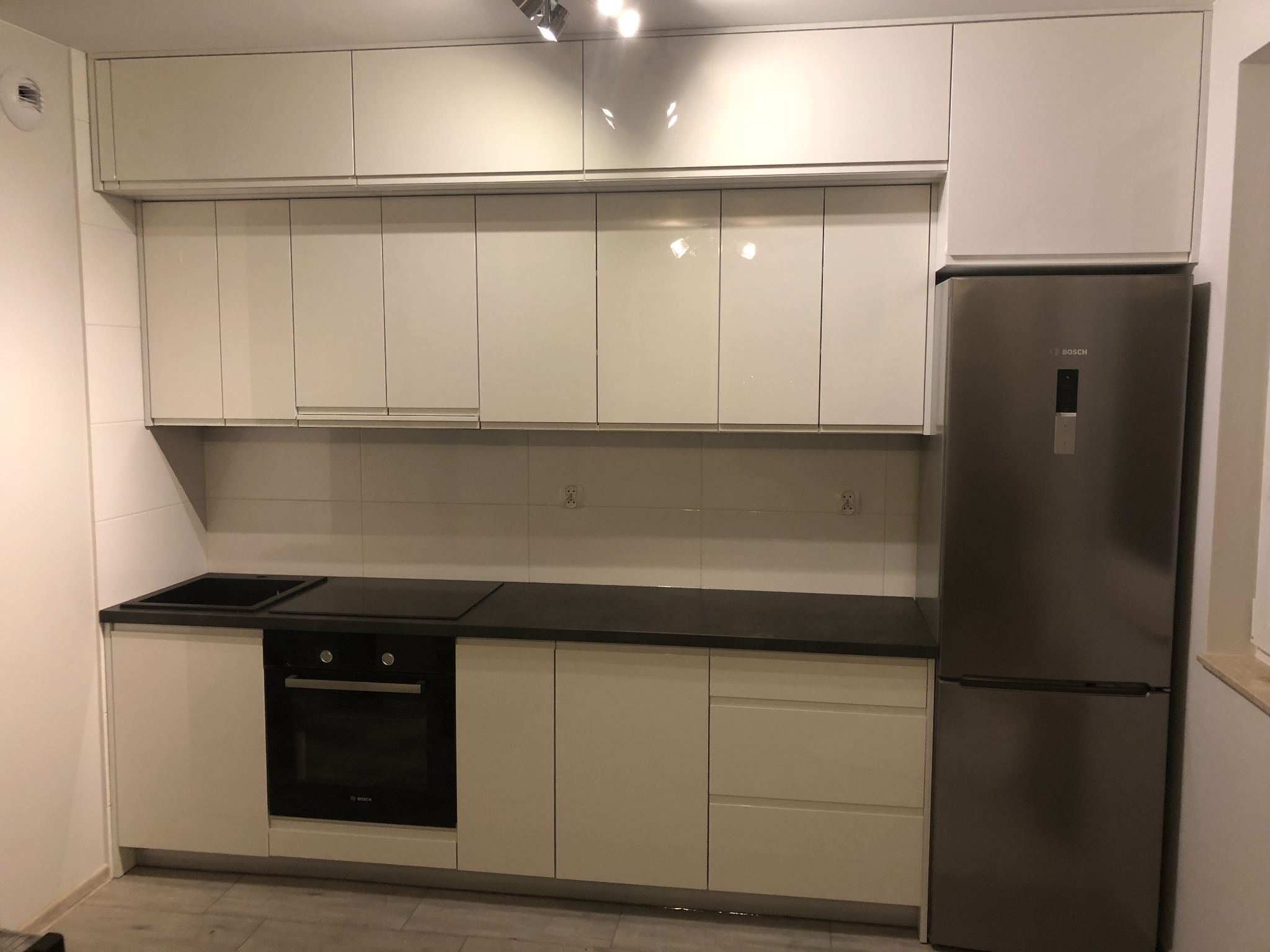 Nowoczesna, biała zabudowa kuchenna z błyszczącymi frontami, czarnym blatem i srebrną lodówką Bosch, oświetlona halogenowymi lampkami.