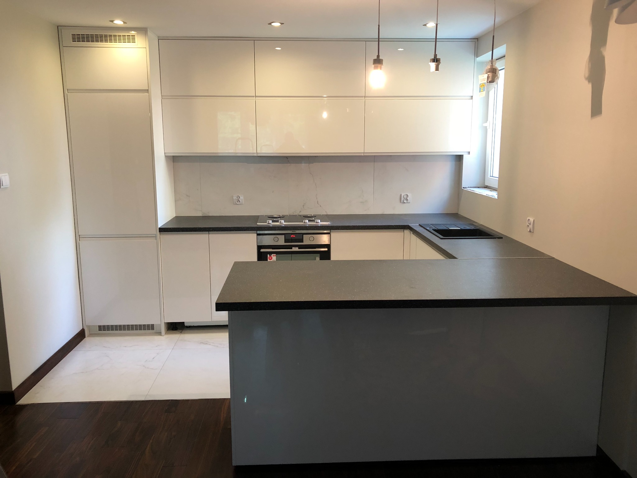 Nowoczesna kuchnia z białymi, lakierowanymi frontami szafek, szarym blatem i wyspą, oświetlona wiszącymi lampami z żarówkami Edisona.