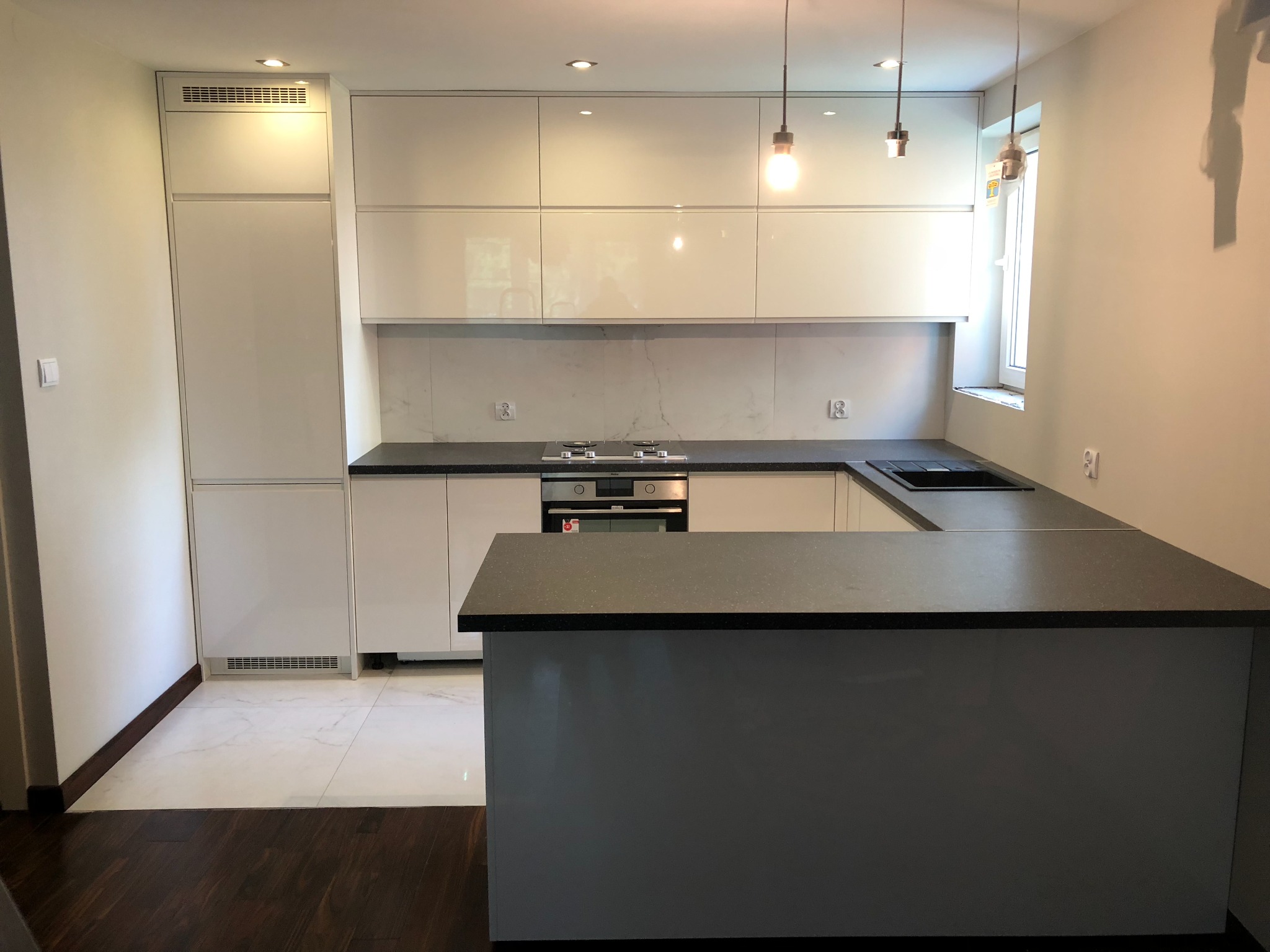 Nowoczesna, jasna kuchnia z wyspą w Karniowicach. Białe, lakierowane fronty szafek, ciemny blat roboczy i marmurowe płytki na ścianie. Nad wyspą wiszą dwie lampy z żarówkami Edisona.