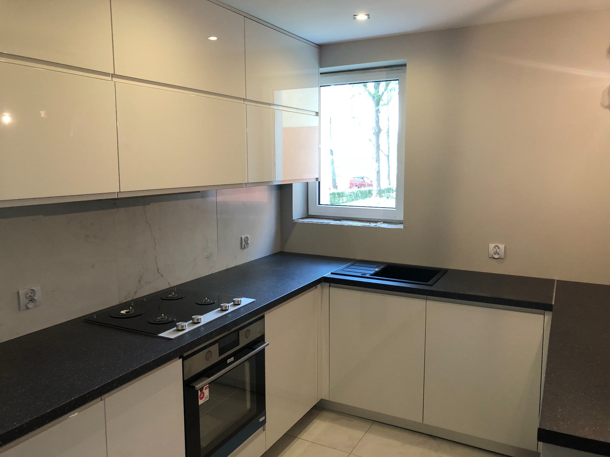 Nowoczesna kuchnia z białymi, lakierowanymi frontami szafek, czarnym blatem z drobinkami i płytą indukcyjną, widok na okno z krajobrazem za szybą.