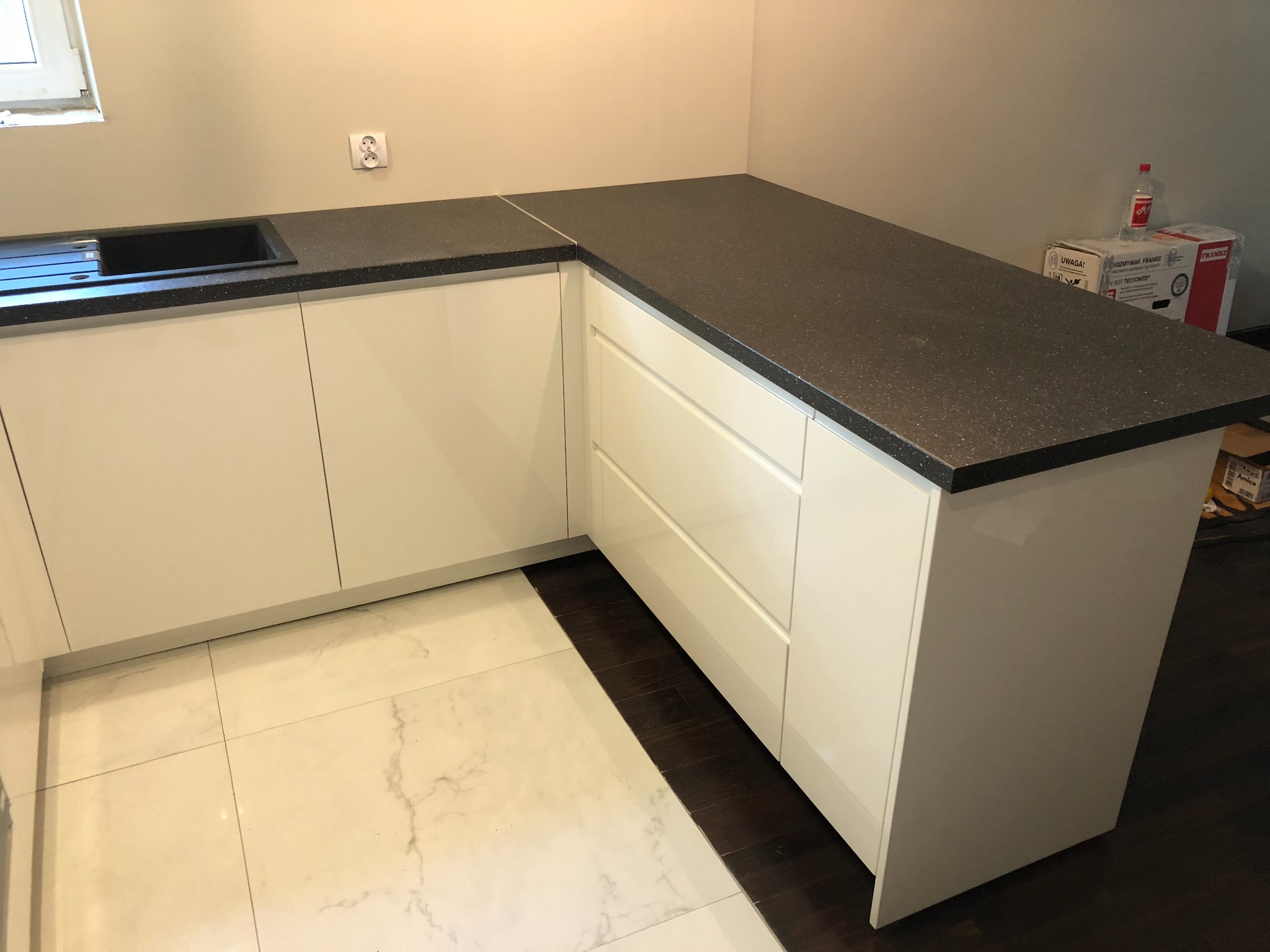 Nowoczesna kuchnia z białymi, lakierowanymi frontami i ciemnym blatem z drobinkami, widoczny zlew i gniazdko elektryczne na ścianie, podłoga wyłożona płytkami imitującymi marmur.