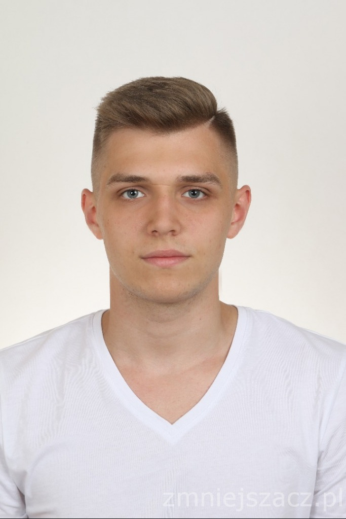 Portret młodego mężczyzny z krótkimi, brązowymi włosami zaczesanymi na bok, ubrany w biały T-shirt z dekoltem w serek, na jasnym tle.