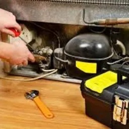 Otwarta lodówka od tyłu, widoczne elementy agregatu. Mężczyzna w trakcie pracy, trzyma śrubokręt w prawej dłoni. Na podłodze leży klucz i czarna skrzynka narzędziowa z żółtymi elementami.