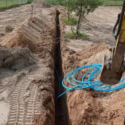 Długi, wąski wykop w ziemi z ułożonym niebieskim przewodem, widoczna łyżka koparki, rów z hałdami ziemi po bokach, w tle drzewa i ogrodzenie.