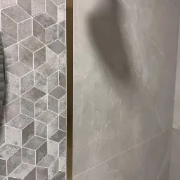 Szare płytki w łazience: geometryczny wzór kostki 3D po lewej stronie, kontrastujące z gładkimi, marmurkowymi płytkami po prawej stronie, oddzielone złotą listwą.