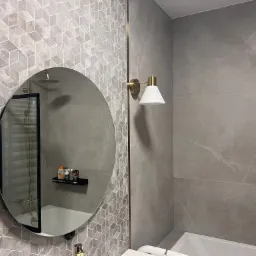 Nowoczesna łazienka w odcieniach szarości z geometryczną mozaiką na ścianie, okrągłe lustro odbija kabinę prysznicową, a złota lampa ścienna dodaje elegancji.