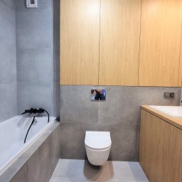 STIM-BUD SP. Z.O.O - Nowoczesna łazienka z szarymi płytkami, drewnianymi szafkami i podwieszaną toaletą. Minimalistyczny design, oświetlenie punktowe, wanna z czarną armaturą.