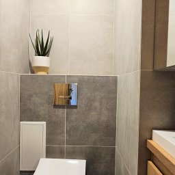 STIM-BUD SP. Z.O.O - Nowoczesna łazienka z szarymi płytkami, chromowanym przyciskiem spłuczki i rośliną w doniczce. Minimalistyczny design, jasne fugi, fragment umywalki z drewnianą szafką.