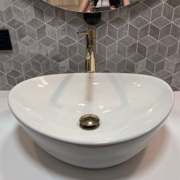 STIM-BUD SPÓŁKA Z OGRANICZONĄ ODPOWIEDZIALNOŚCIĄ - Biała, owalna umywalka nablatowa z złotą armaturą na tle szarej ściany z geometrycznym wzorem. Widoczne odbicie w lustrze.