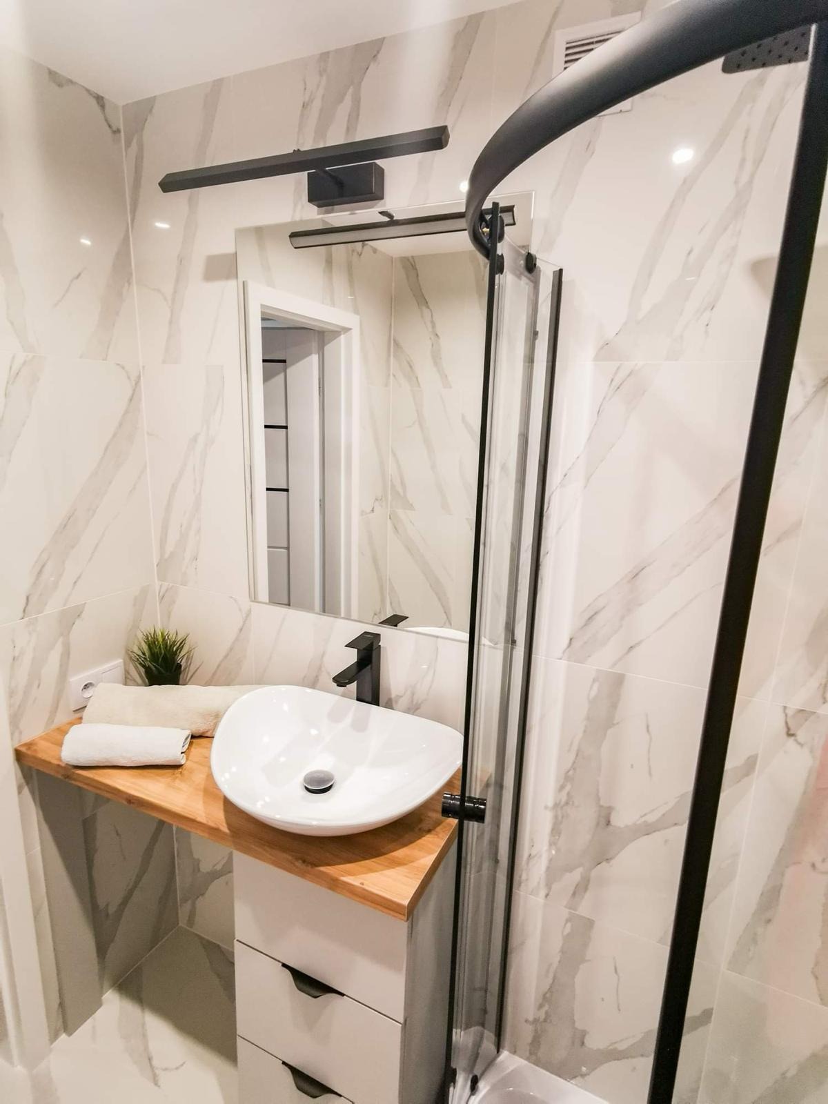 Nowoczesna łazienka z marmurowymi płytkami, białą umywalką nablatową na drewnianym blacie, czarną armaturą i kabiną prysznicową z czarnymi profilami.