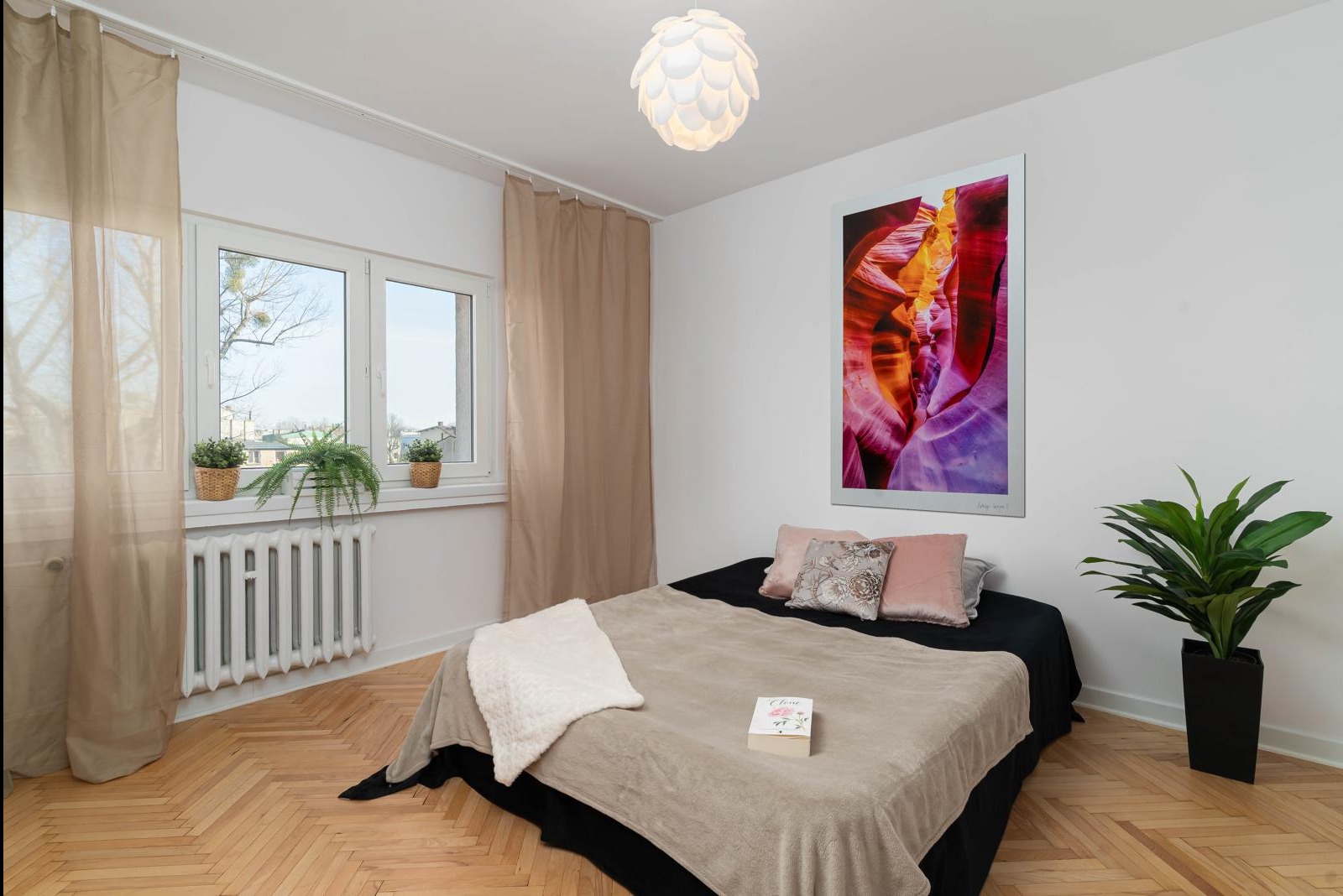 Jasna sypialnia z parkietem w jodełkę, beżowymi zasłonami, łóżkiem z beżową narzutą, kolorowym obrazem na ścianie i roślinami doniczkowymi.