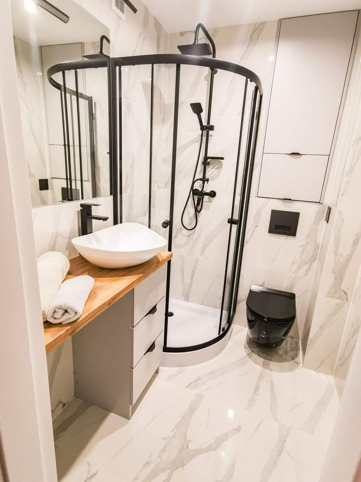 Nowoczesna łazienka z kabiną prysznicową z czarnymi profilami, białą umywalką na drewnianym blacie, czarną armaturą i toaletą w kolorze czarnym, marmurowe płytki na ścianach i podłodze.