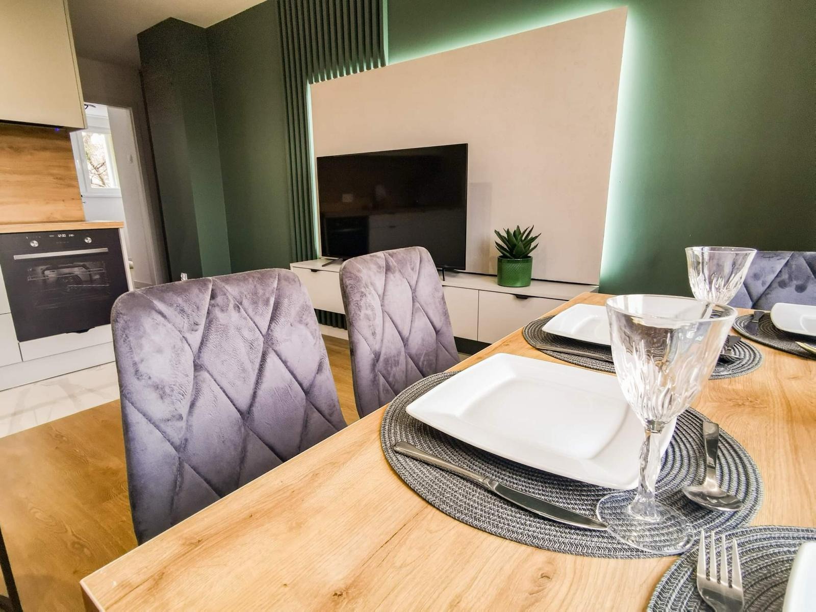 Nowoczesna jadalnia z drewnianym stołem, nakrytym dla dwóch osób, z szarymi, welurowymi krzesłami i zielonym akcentem dekoracyjnym na ścianie z podświetleniem LED.