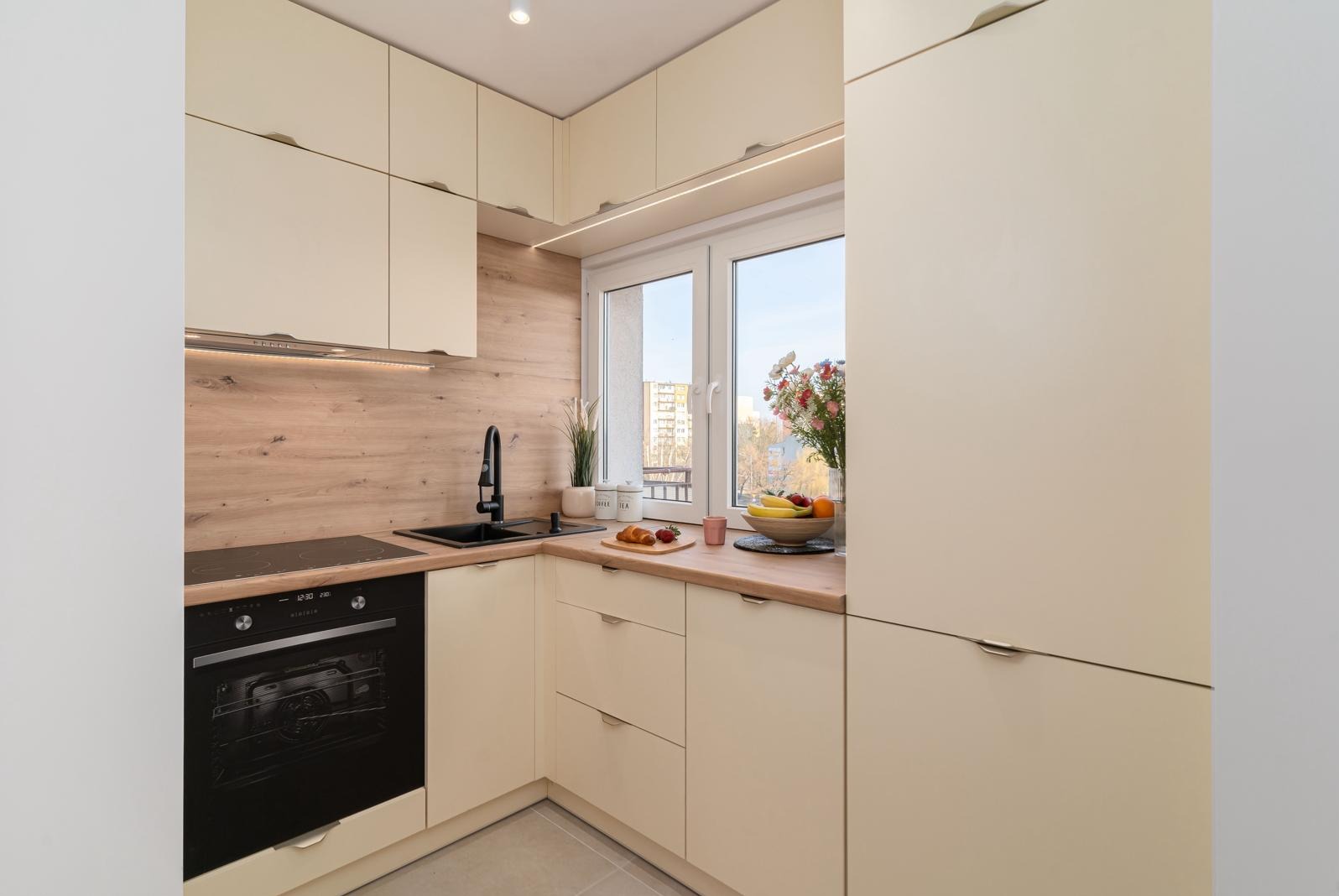 Jasna kuchnia z drewnianym panelem, czarną armaturą i owocami na blacie przy oknie.