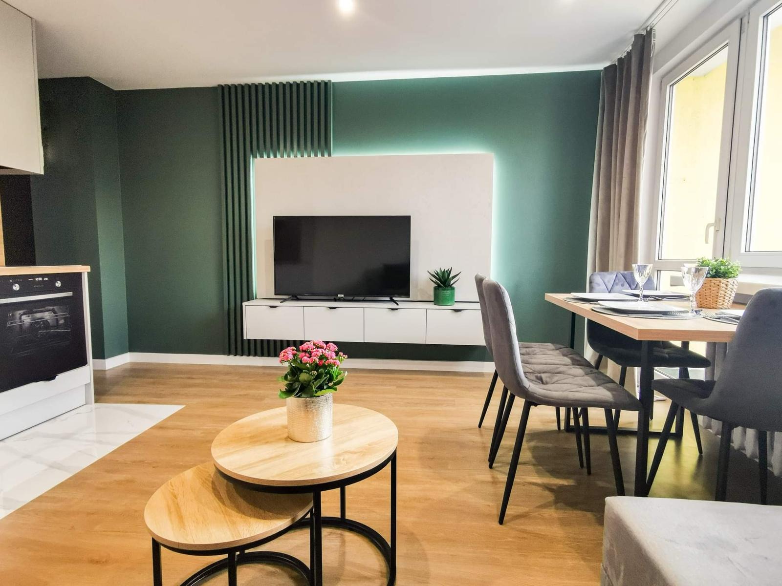 Nowoczesne wnętrze z zieloną ścianą, podświetlanym panelem TV, drewnianą podłogą i stolikiem kawowym z kwiatami.