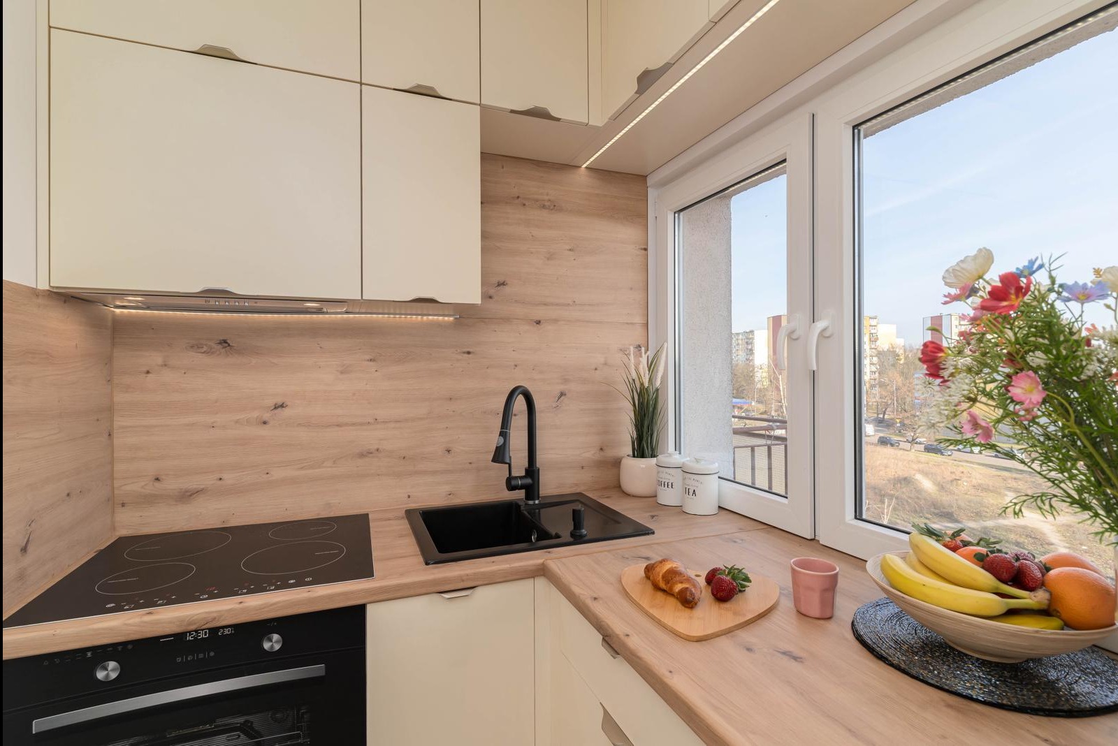 Nowoczesna kuchnia z drewnianymi panelami ściennymi, czarnym zlewem i płytą indukcyjną, oświetleniem LED pod szafkami, oraz dekoracyjnymi elementami jak owoce, kwiaty i pojemniki na kawę i herbatę.