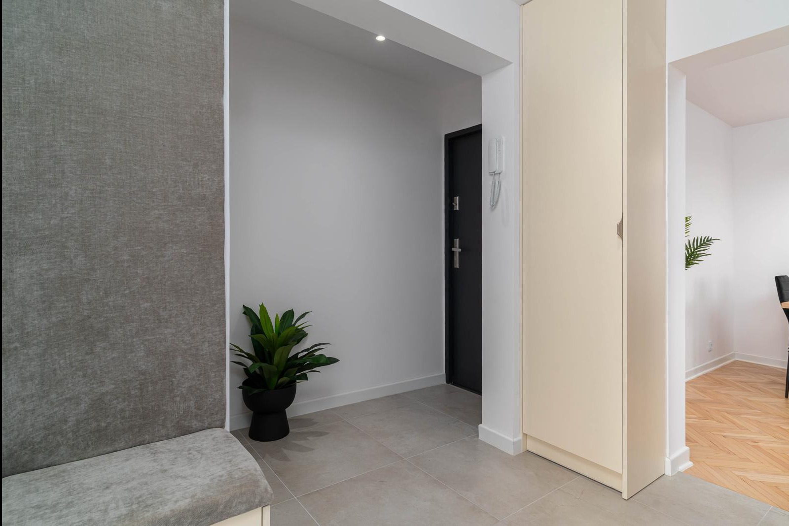 Nowoczesny przedpokój z szarą tapicerowaną ścianą, czarnymi drzwiami wejściowymi, beżową szafą wnękową i rośliną doniczkową w czarnej osłonce, wykończony płytkami podłogowymi w odcieniach szarości.