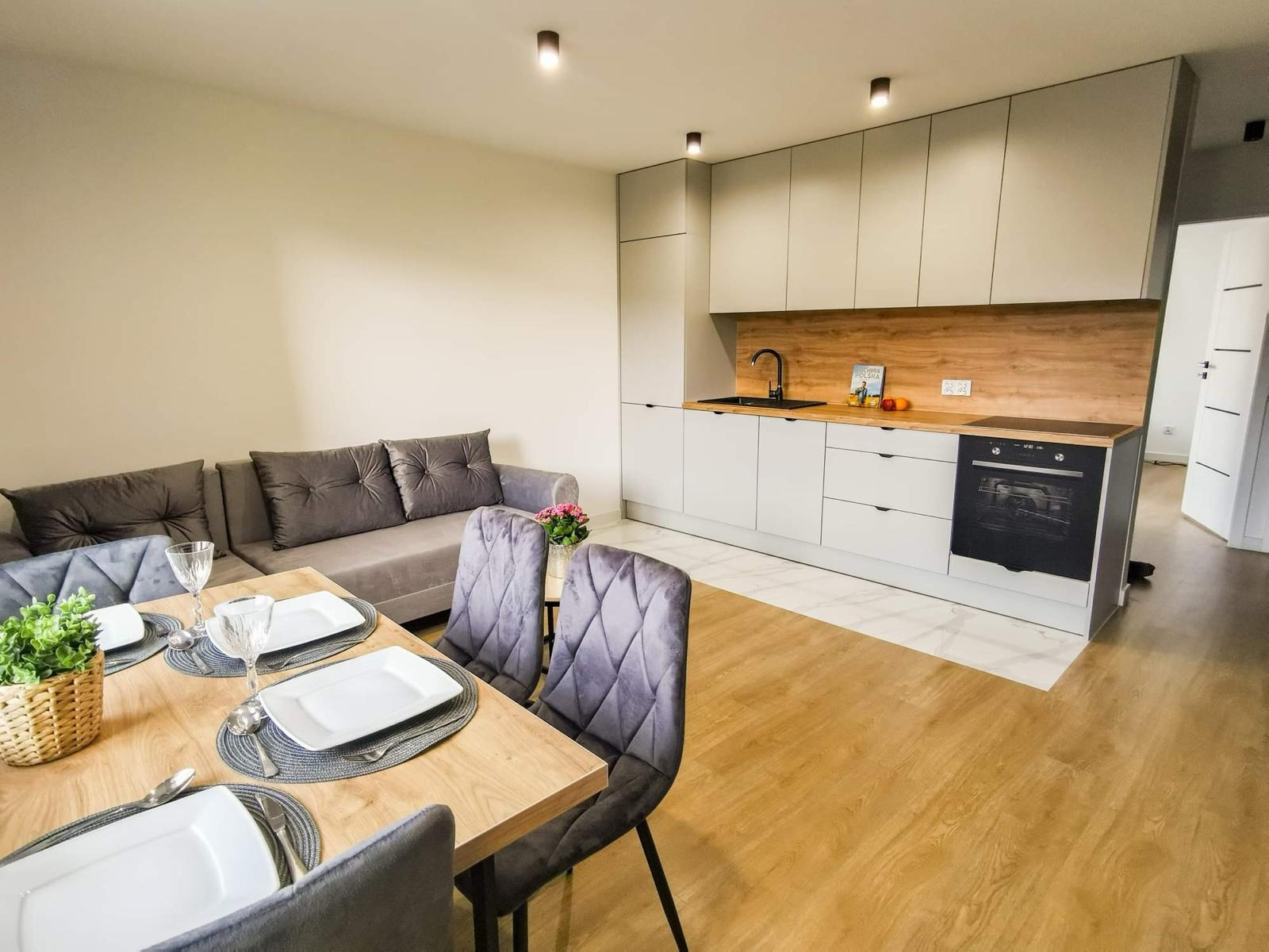 Jasne wnętrze mieszkania po remoncie: stół z nakryciem, szare krzesła, sofa z poduszkami i nowoczesna zabudowa kuchenna z drewnianym blatem i czarną armaturą.
