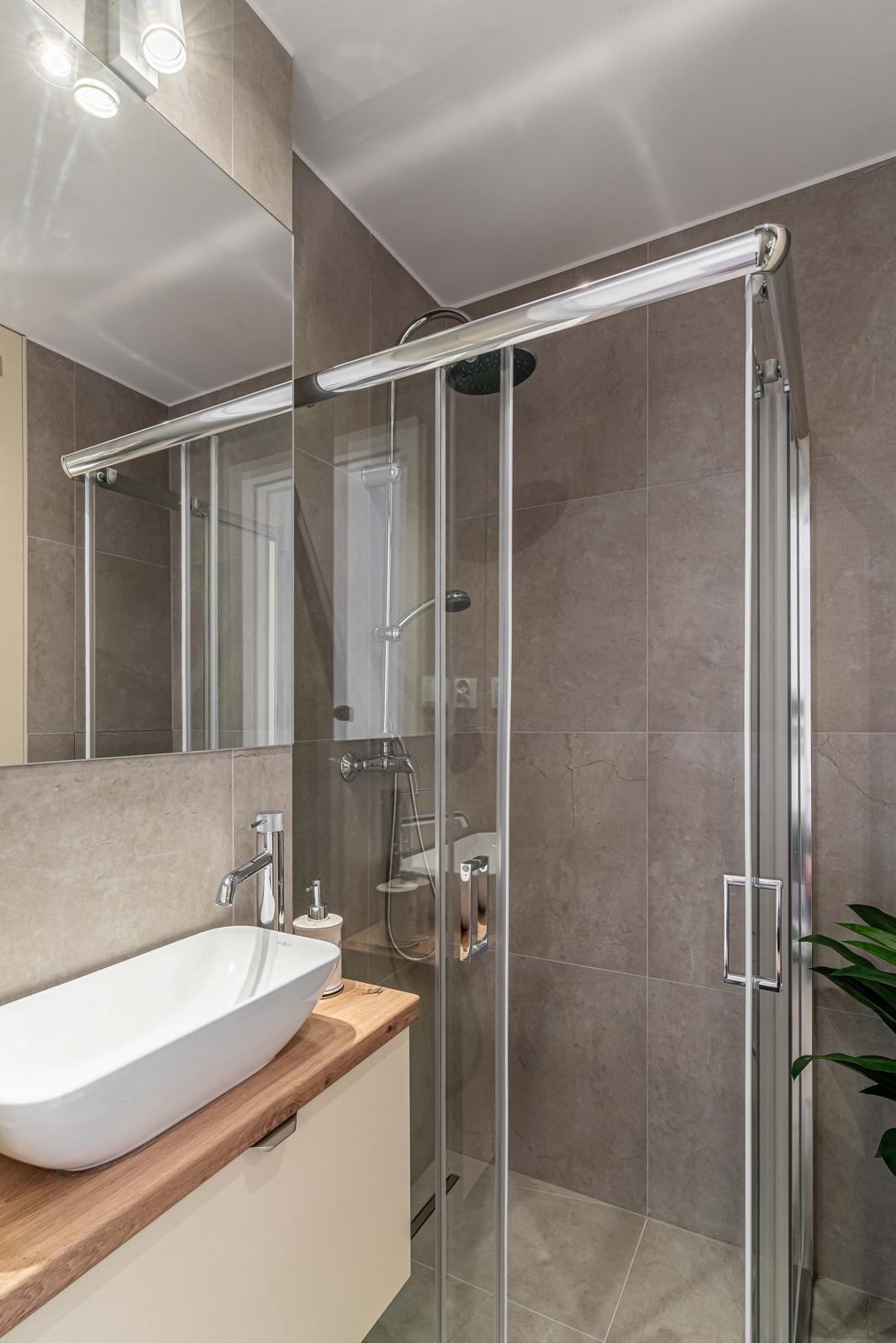 Nowoczesna łazienka z kabiną prysznicową ze szklanymi drzwiami przesuwnymi, umywalką nablatową o owalnym kształcie na drewnianym blacie i szare płytki imitujące kamień na ścianach.