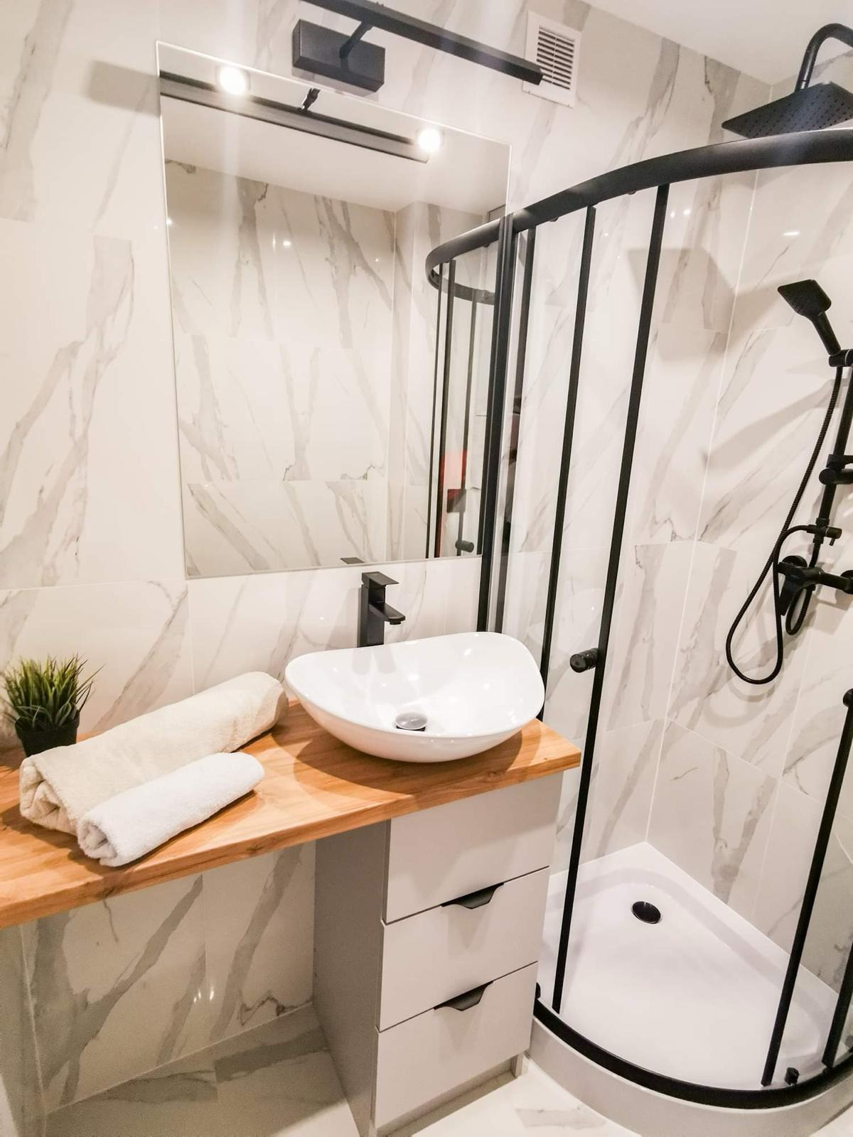 Nowoczesna łazienka z kabiną prysznicową w czarnej ramie, umywalką nablatową na drewnianym blacie i lustrem z oświetleniem punktowym.