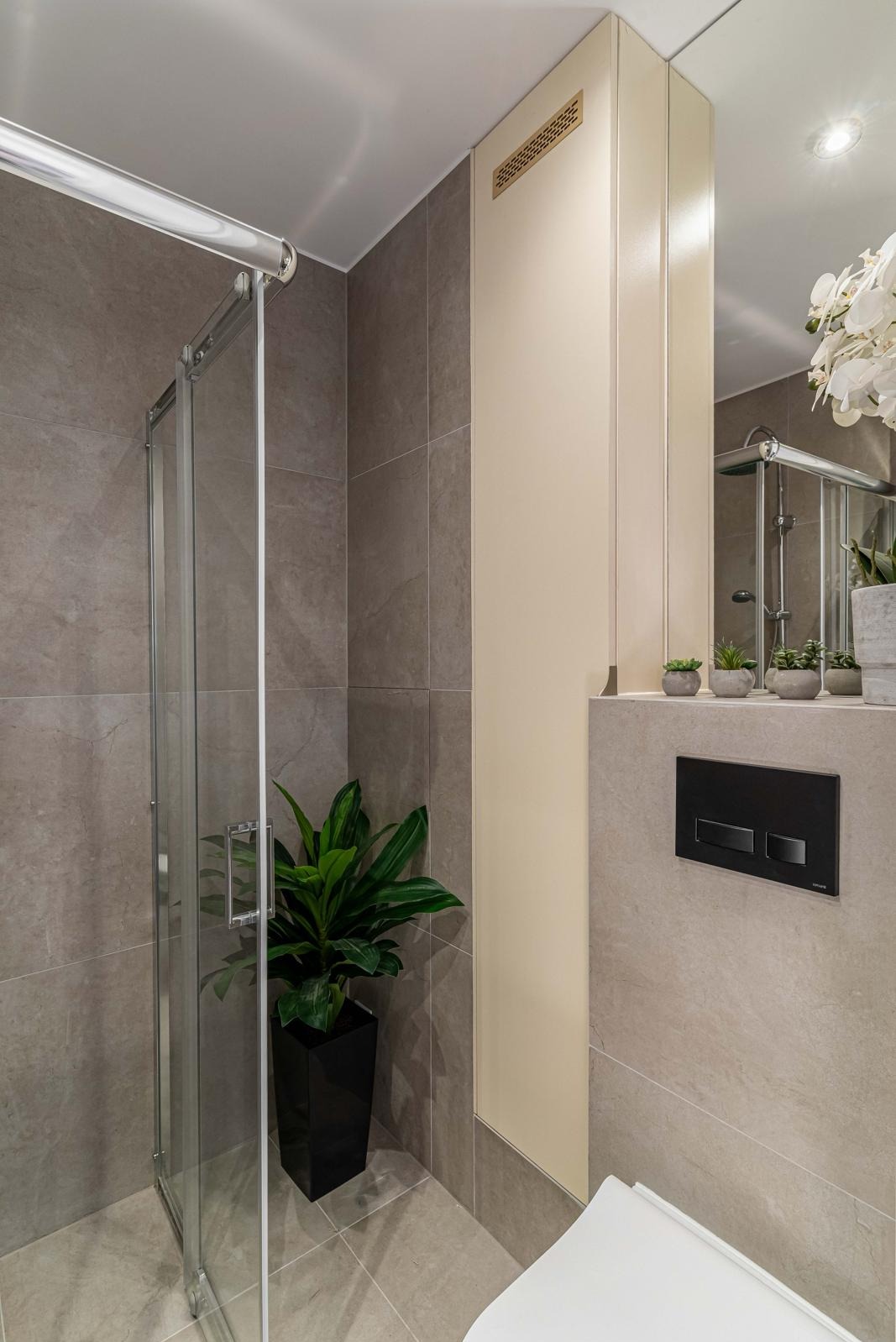 Nowoczesna łazienka z kabiną prysznicową ze szklanymi drzwiami przesuwnymi, szare płytki wielkoformatowe, zabudowa w kolorze kremowym z kratką wentylacyjną, czarny panel sterujący spłuczką, roślina...