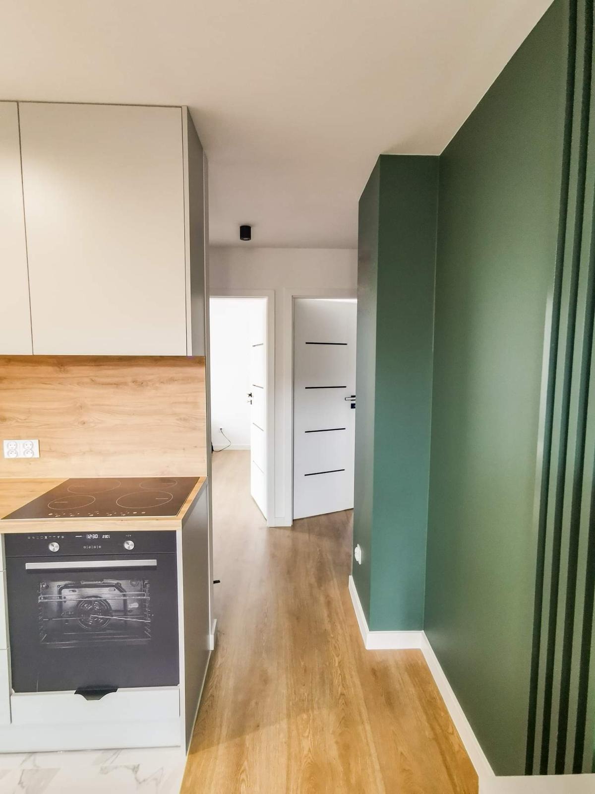 Nowoczesna kuchnia z elementami drewna, widok na przedpokój z białymi drzwiami z czarnymi paskami, ściana pomalowana na ciemny zielony kolor z pionowymi czarnymi listwami.