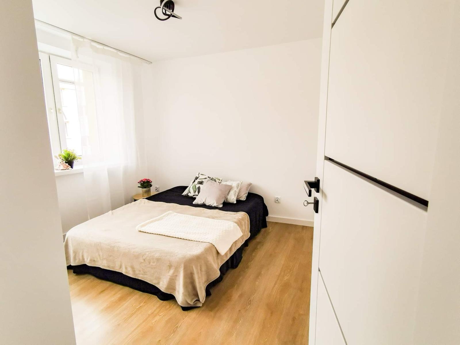 Jasna sypialnia z łóżkiem przy oknie, drewniana podłoga i białe ściany, widok zza uchylonych białych drzwi z czarnymi akcentami.