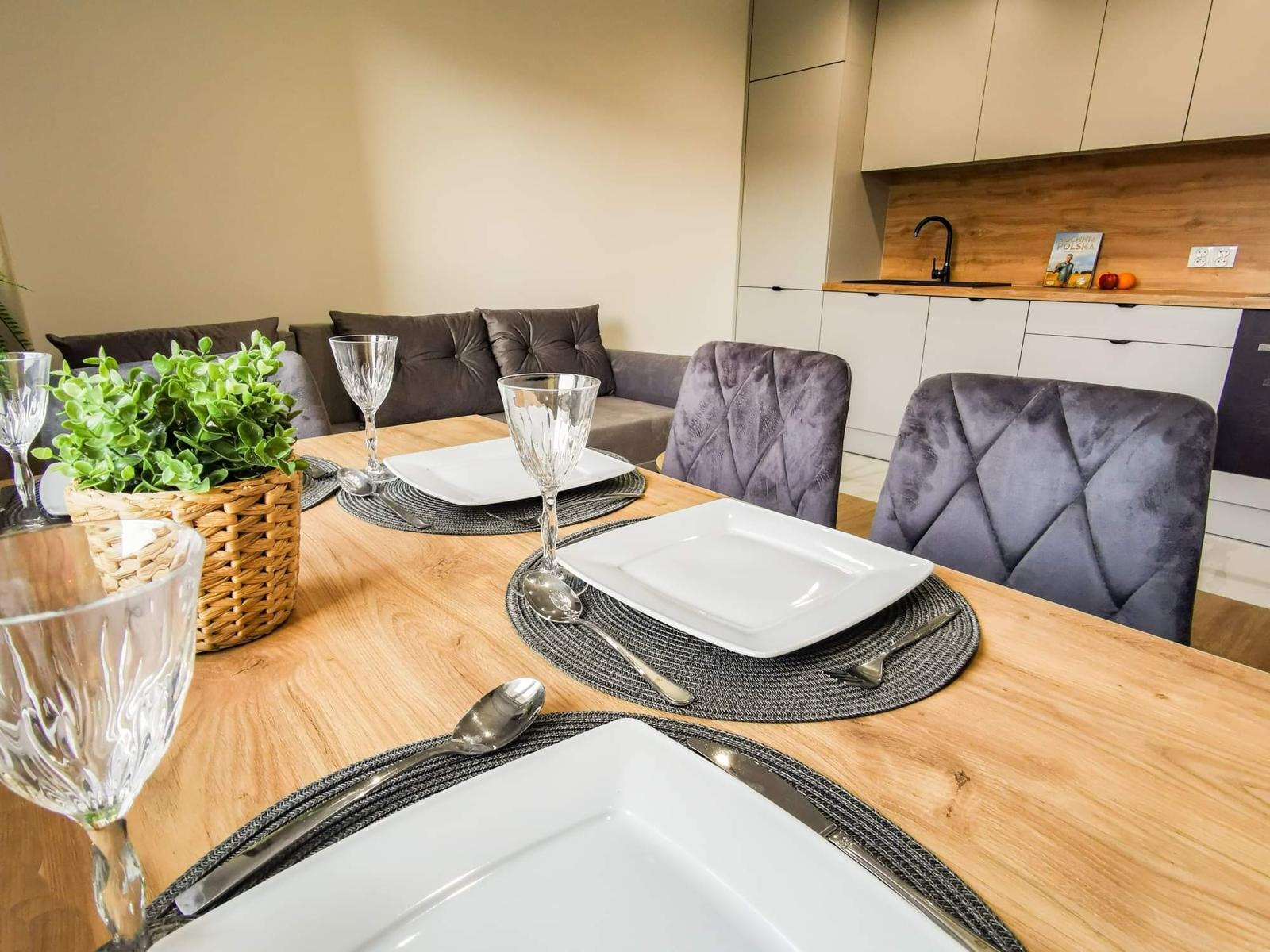 Stół z jasnego drewna, nakryty dla trzech osób białymi, kwadratowymi talerzami na szarych podkładkach, srebrne sztućce, kryształowe kieliszki, w tle szara kanapa i zabudowa kuchenna z drewnianym...
