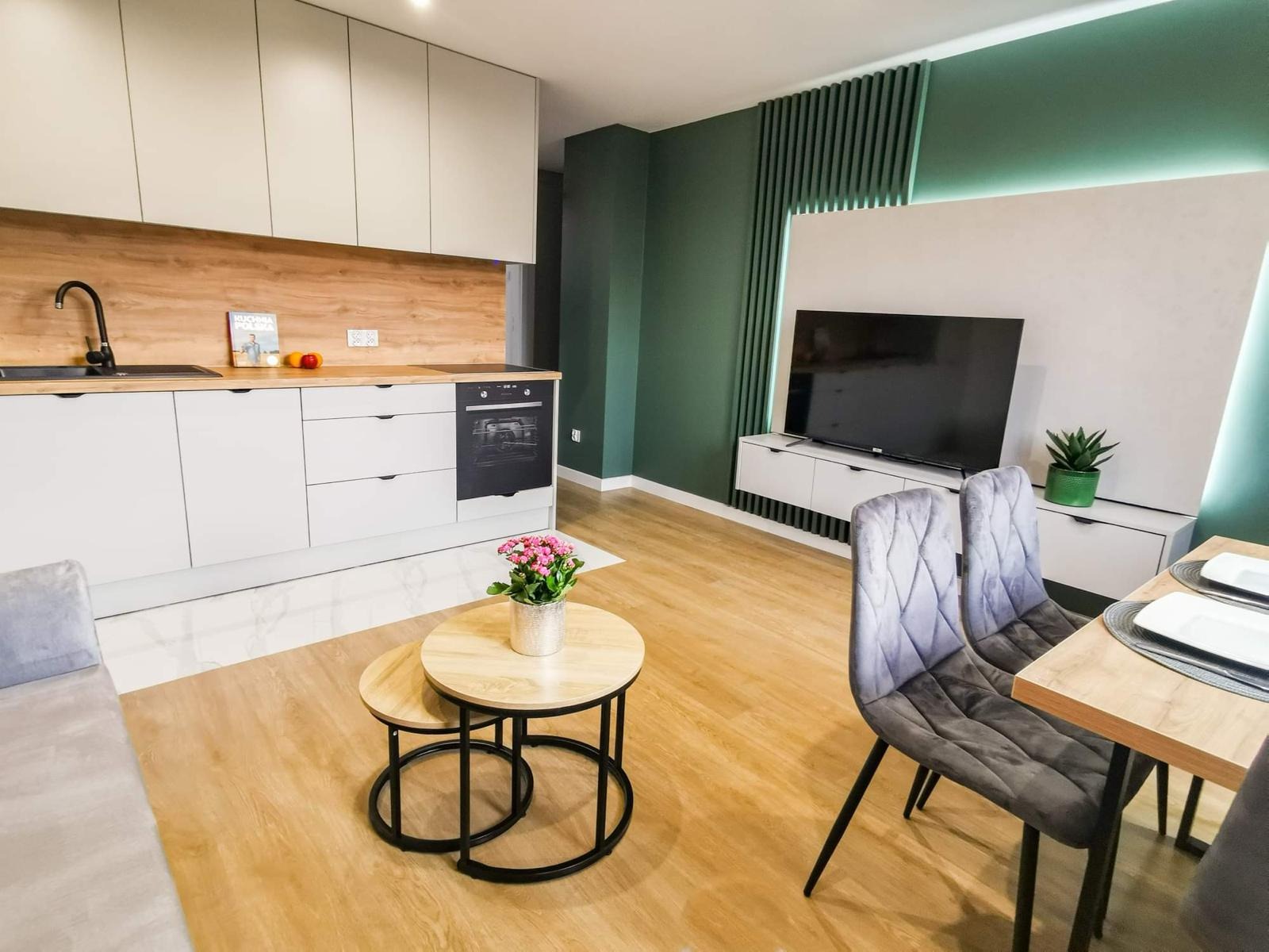Jasna kuchnia z drewnianym blatem przechodząca w salon z zieloną ścianą akcentową i telewizorem, widoczne krzesła z szarego weluru przy stole.