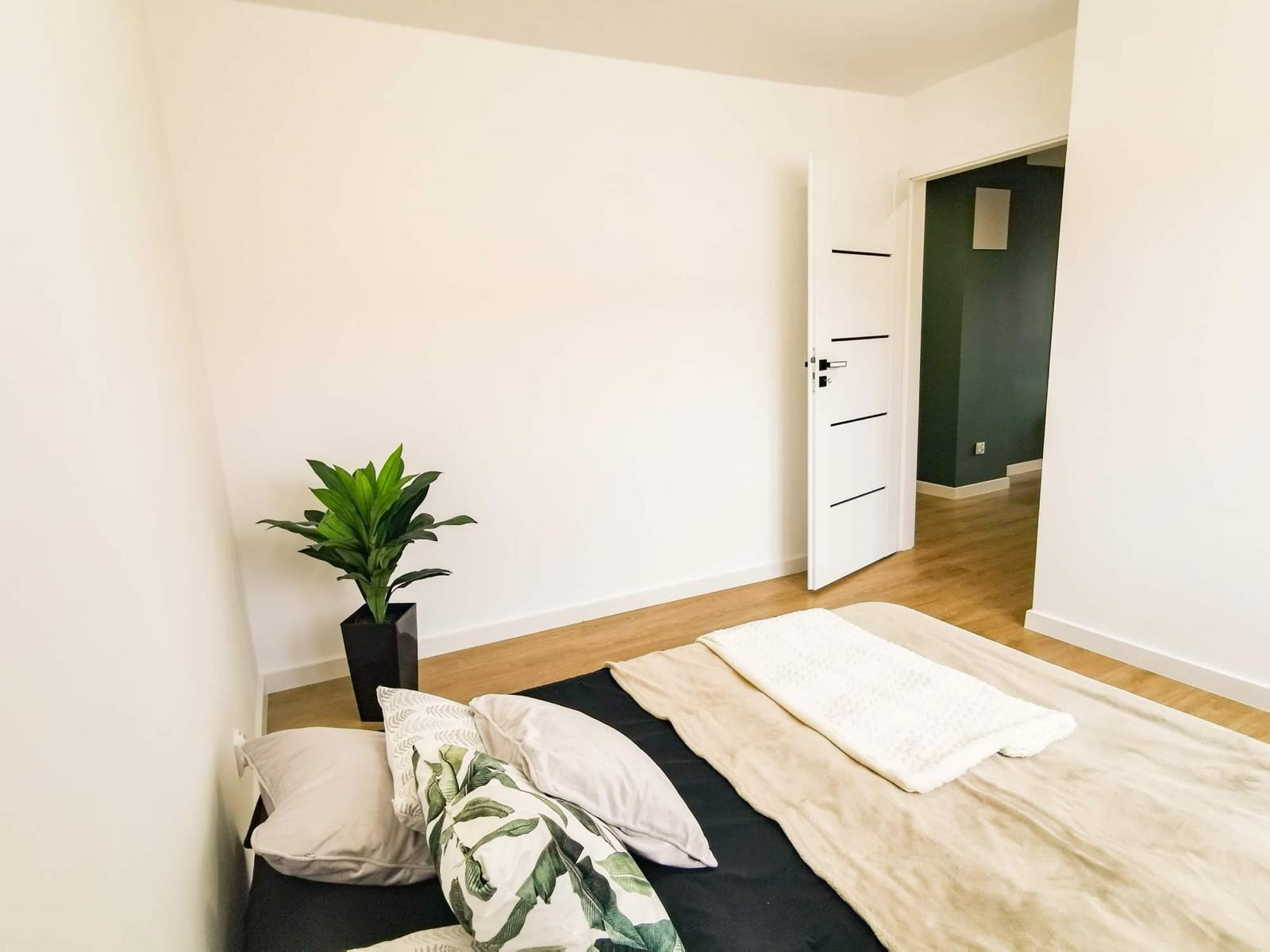 Jasna sypialnia z drewnianą podłogą, białymi ścianami i otwartymi białymi drzwiami z czarnymi akcentami, prowadzącymi do ciemnozielonego pokoju; na pierwszym planie łóżko z czarną pościelą...