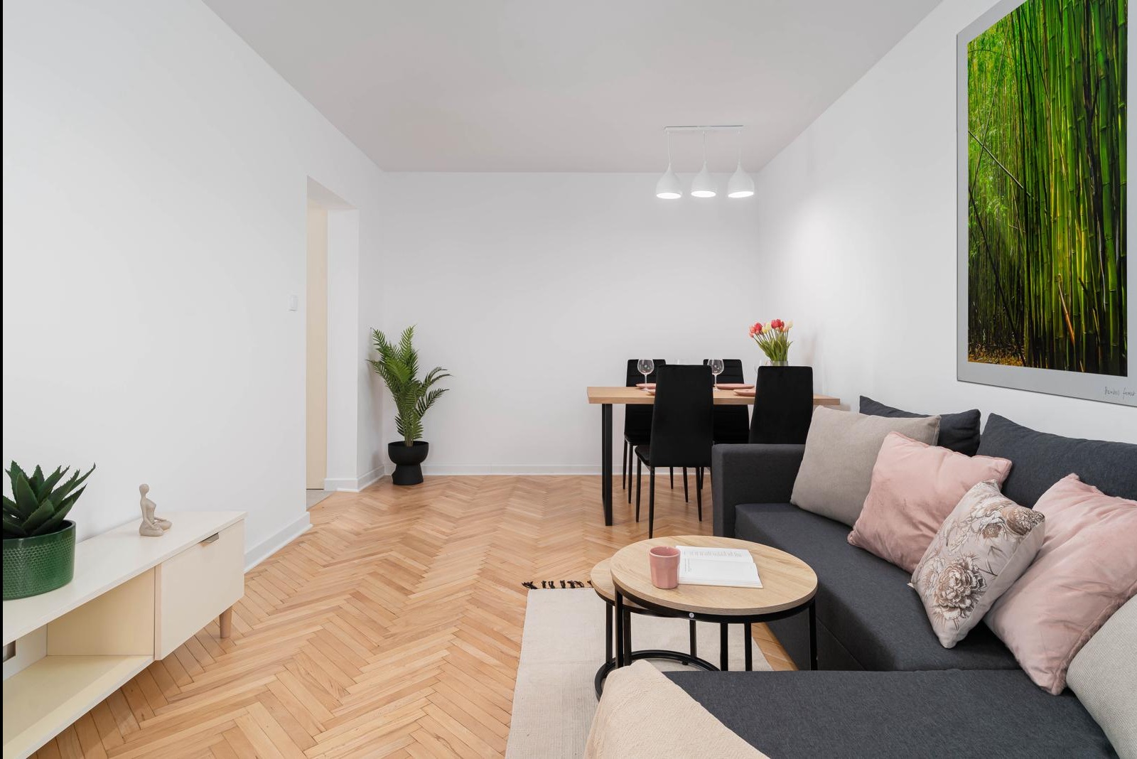 Jasne, przestronne wnętrze po remoncie: parkiet w jodełkę, szara sofa z różowymi poduszkami, stół z czarnymi krzesłami, minimalistyczne oświetlenie i grafika z motywem bambusa na ścianie.