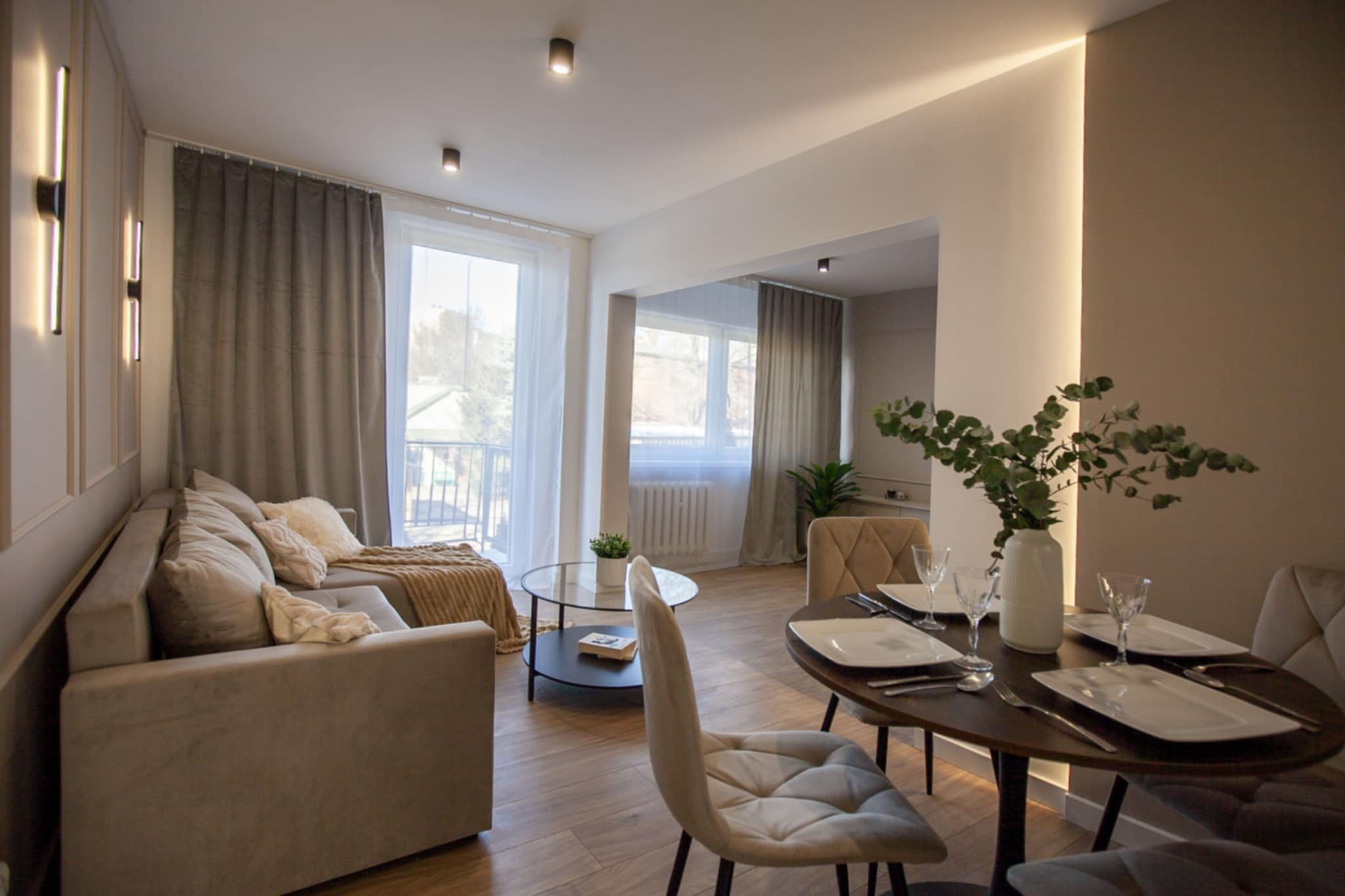 Jasne wnętrze salonu z jadalnią, sofa z poduszkami, okrągły stolik kawowy z książką, stół jadalny z nakryciem dla czterech osób i wazonem z zielonymi gałązkami, podłoga wyłożona panelami w kolorze...