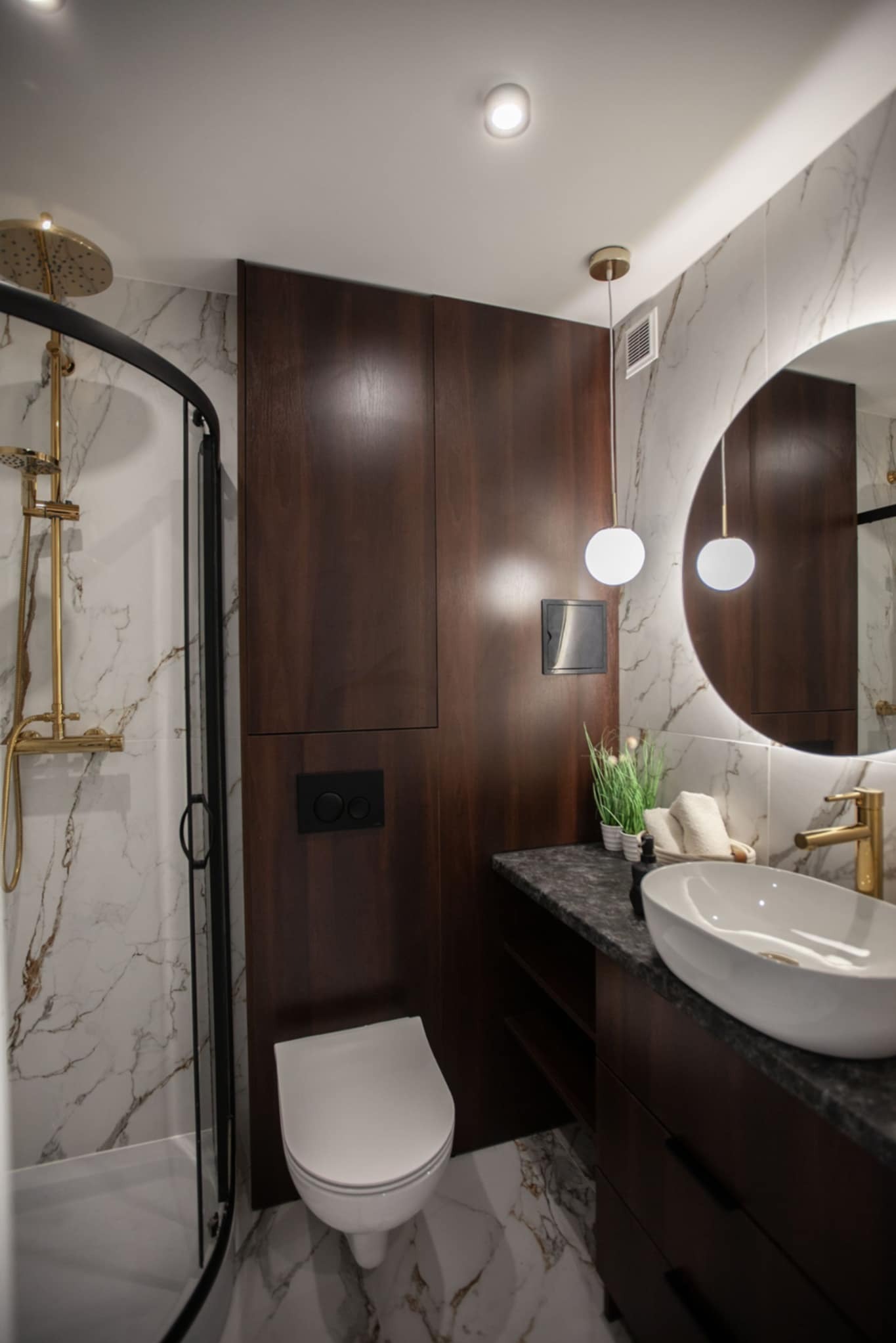 Nowoczesna łazienka z kabiną prysznicową, umywalką nablatową i podwieszaną toaletą, marmurowe płytki na ścianach i podłodze, ciemne drewniane szafki, okrągłe lustro z podświetleniem LED i złota...