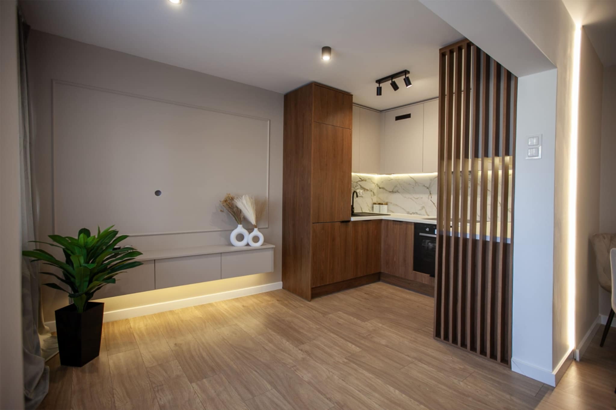 Nowoczesne wnętrze: drewniana podłoga, beżowe ściany z listwami, zabudowa kuchenna z marmurowym blatem, drewniana zabudowa z oświetleniem LED pod spodem, dekoracyjna ścianka z drewnianych listew.